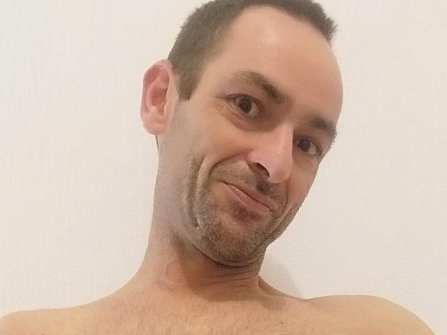Ledjee has a balding head, short dark hair, and a light beard with a slight smirk, showing his shirtless upper body. His brown eyes and light skin are visible.