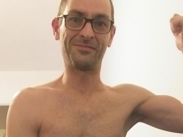Ledjee, shirtless, with glasses, light skin, short brown hair, slight beard, smiling, upper body, right arm raised, chest visible, medium build, slight chest hair, casual selfie.