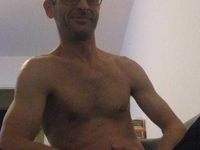 Shirtless Ledjee with a muscular, toned torso, slight chest hair, small nipples, and a subtle smile, wearing glasses, sitting indoors.