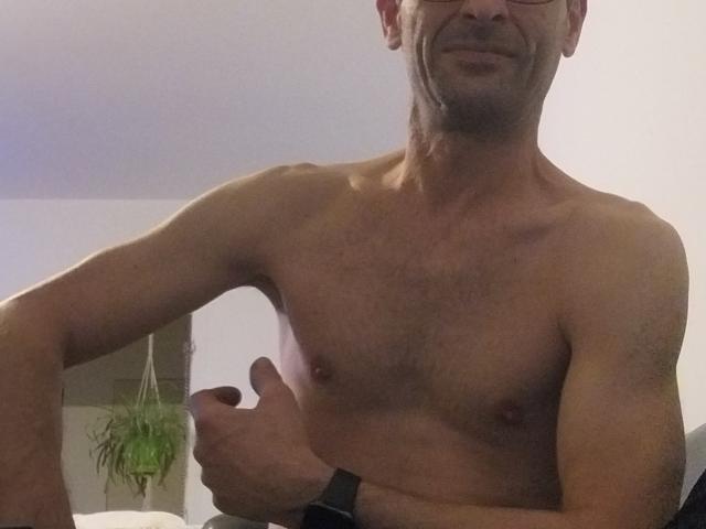 Shirtless Ledjee with a muscular, hairy chest and a thumbs-up, wearing glasses and a watch, smiling slightly. His skin is tan, and he's in a dimly lit room.