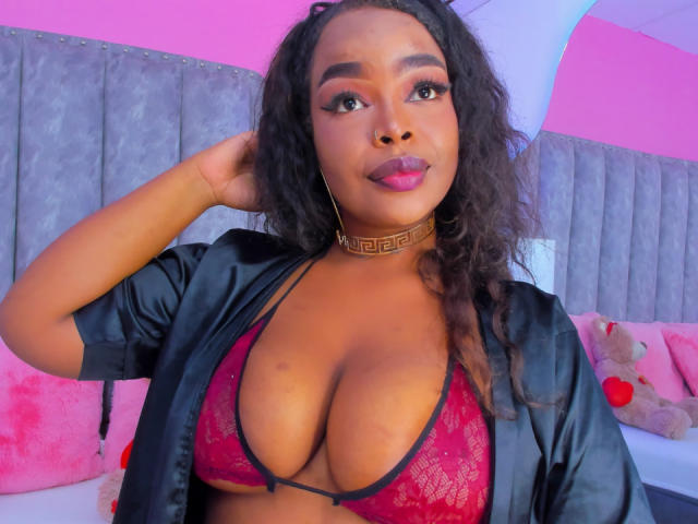 MadinsonLopez with dark curly hair, wearing a red lace bra that shows her big tits, black satin robe, gold choker, and pink lipstick, looking at the camera.