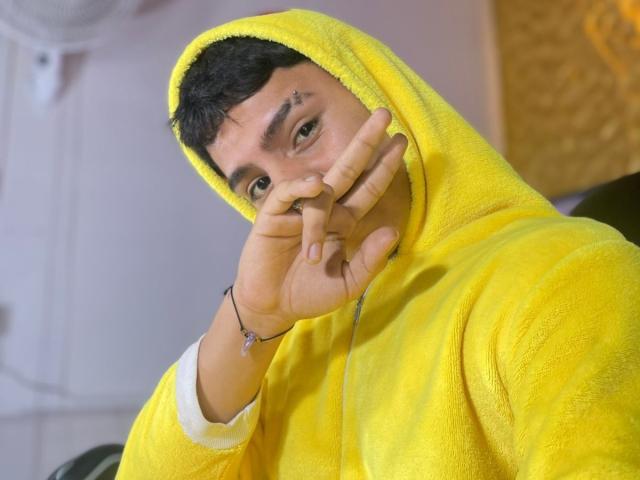 RichiMontes, vistiendo una sudadera suave de color amarillo brillante con la capucha puesta, se tapa la boca con la mano, mostrando un piercing de metal en la nariz y una pulsera con un pequeño dije..