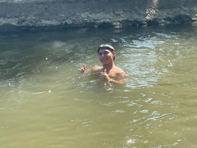 RichiMontes, con el cabello oscuro y mojado, está en agua poco profunda de color verdoso, mostrando una pequeña sonrisa y levantando el pulgar con su mano derecha. Está sin camisa, con una complexión delgada..