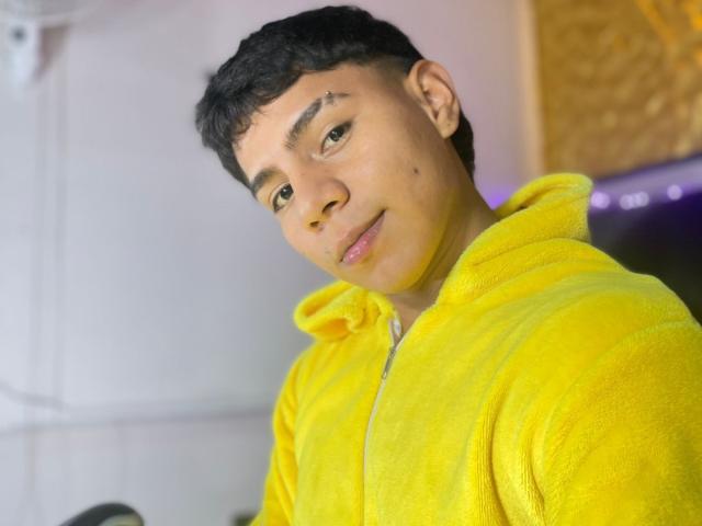 RichiMontes, die een felgele hoodie draagt, heeft kort zwart haar, een kleine wenkbrauw tattoo en een lichte glimlach, met een schattige, ontspannen uitdrukking en lichtbruine huid..