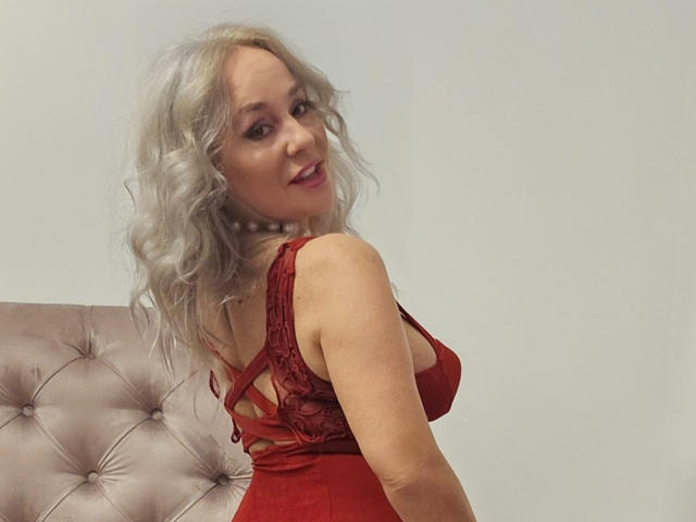 EmilyLowe69 with silver curly hair, red lace dress, showing her tits, looking over her shoulder, smiling, standing in front of a tufted beige headboard.