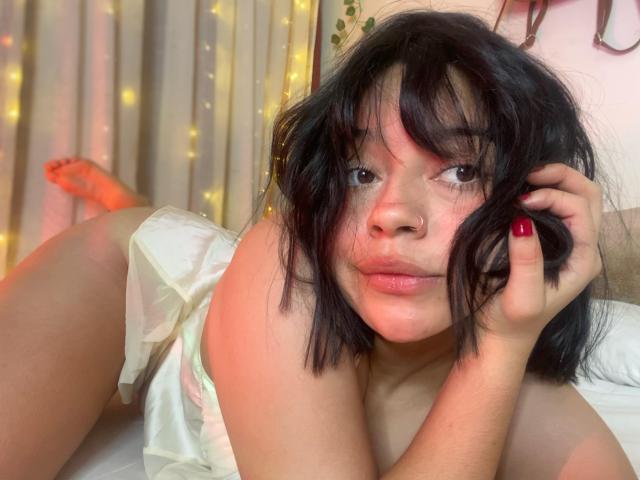 CuteSara with messy black hair, pink nose, and red nails, lying on a bed in white lingerie, looking at the camera with a slight pout, legs up.