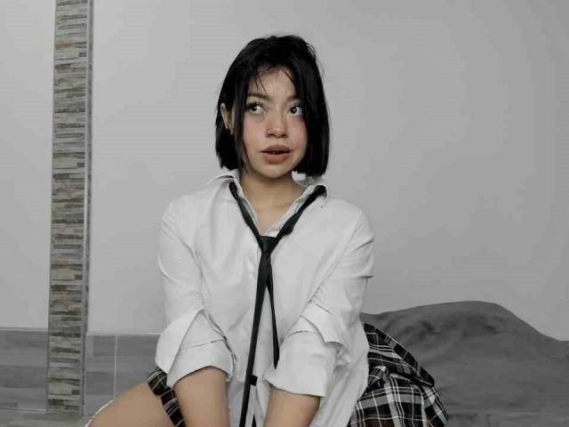 CuteSara with short black hair, wearing a white shirt and black tie, sitting on a bed, plaid skirt, surprised expression, looking at camera, hands on knees.
