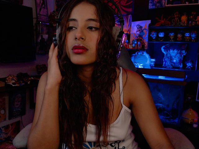 RedTemptation with wet brown hair, wearing a white tank top, sitting with large headphones on, red lipstick, looking slightly to the side, hand touching ear.
