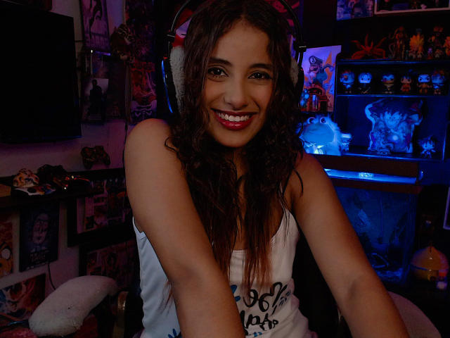 RedTemptation has curly brown hair, tan skin, and red lipstick, smiling with white tank top, showing her tits, in dimly lit room with blue and purple neon lights.