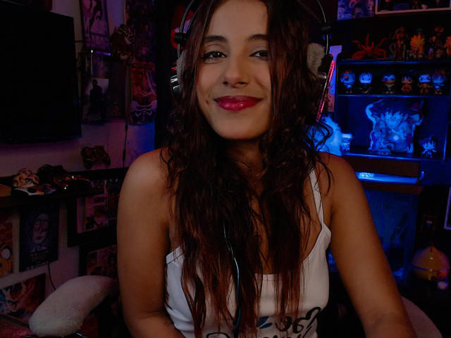 RedTemptation with wavy brown hair, wearing headphones, smiling with pink lipstick, in a white tank top, showing her tits, in a dimly lit room, surrounded by blue and purple lights.