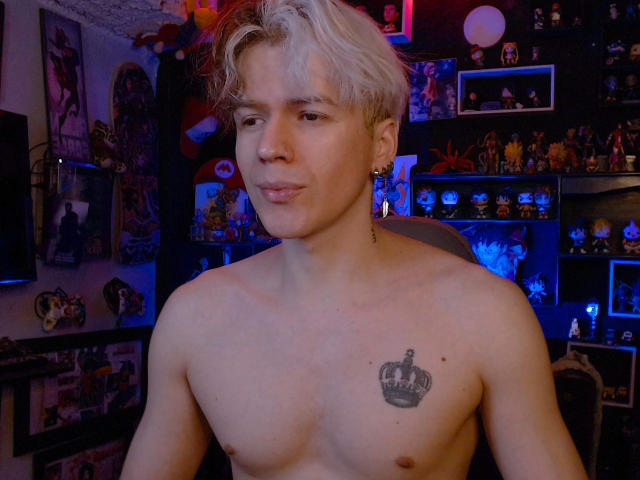 Shirtless RedTemptation with blonde hair, nose ring, and crown tattoo on chest, looking at camera with slight smirk, in dimly lit room with colorful decorations and blue lights.