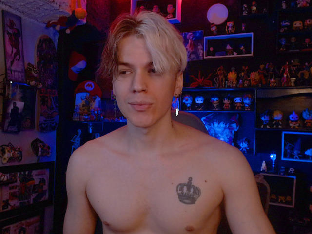 Shirtless RedTemptation with blond and brown hair, small crown tattoo on chest, subtle smile, and blue earrings, standing in dimly lit room filled with posters and figurines.