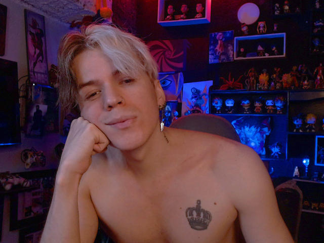 Shirtless RedTemptation with blonde and brown hair, crown tattoo on chest, earings, and a smug expression, resting head on hand, in dimly lit room.