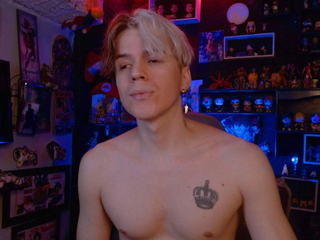 Shirtless RedTemptation with blonde hair, small tattoo of a crown on chest, slight smile, and earrings, standing in dimly lit room filled with colorful toys and decorations.