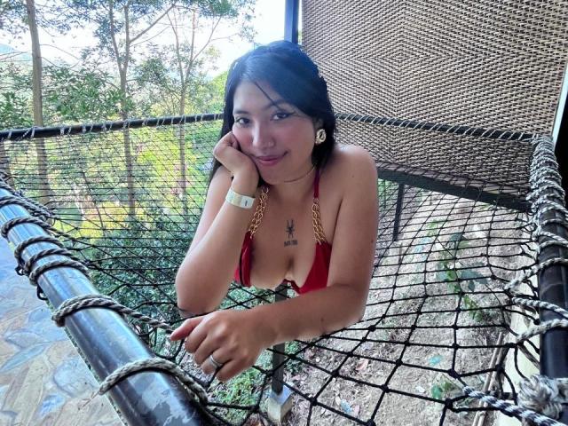 Elizaa, con pechos pequeños, un top de bikini rojo y una cadena dorada, apoyada en una red, sonriendo, cabello oscuro, piel bronceada, reloj de pulsera, fondo de árboles, hamaca con red, día, bosque..