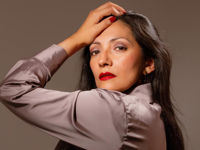 SexyLatinMadura with red lipstick, brown eyes, and long dark hair, wearing a shiny gray satin shirt, hand in hair, looking at camera, brown skin, slightly flushed cheeks.