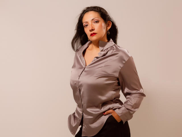SexyLatinMadura with dark hair, red lipstick, and a shiny grey button-up shirt, hands in black pants, standing against a plain wall, looking confident with a slight smirk.