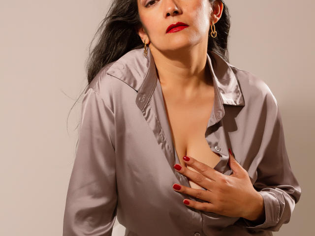 SexyLatinMadura with red lipstick, wearing a shiny grey blouse, hand covering her tits, long black hair, gold earrings, and red nails, looking down with a slightly open mouth.