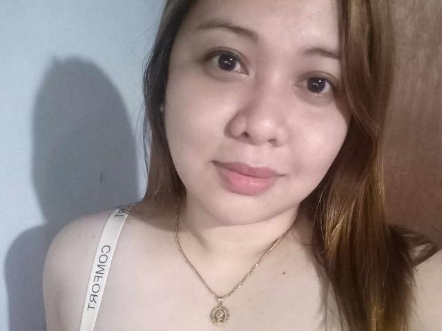 HannaSexy69 has fair skin, brown eyes, and light brown hair. She's wearing a white bra with "THEO MOO" and a gold necklace with a circular pendant. Her lips are slightly parted.