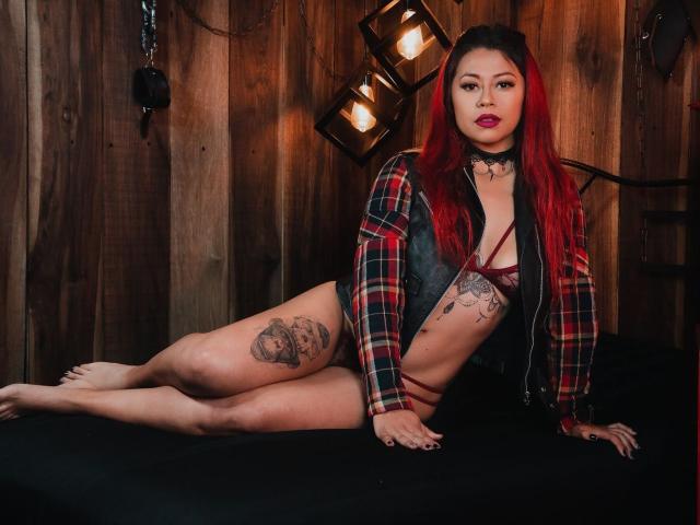 DakotaCrabs with red hair, plaid jacket, black choker, maroon lingerie, tattoos, and a chibi face, leaning on a black bed, showing her tits and a tattooed thigh.