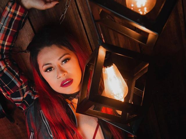 DakotaCrabs with red hair, black choker, and red lipstick, lying under a lantern with a glowing bulb, wearing a plaid shirt and a grey jacket. Her tits are visible.
