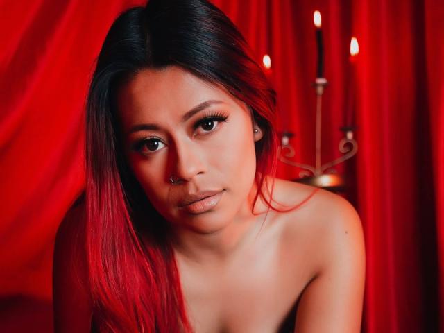 DakotaCrabs with red-tipped black hair, nose ring, and glossy lips, topless, showing her bare shoulders and slightly parted full lips. Red candleholder in the background.