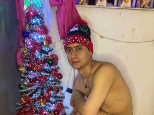 SebastianB69, shirtless with a red and white beanie, sits next to a colorful Christmas tree, arms crossed, wearing a necklace with a cross.