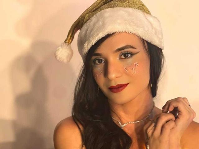 PamelaKander with long dark hair, wearing a gold and white Santa hat, has glitter on her cheeks, red lipstick, and a diamond necklace, holding her hair with a cute smile.
