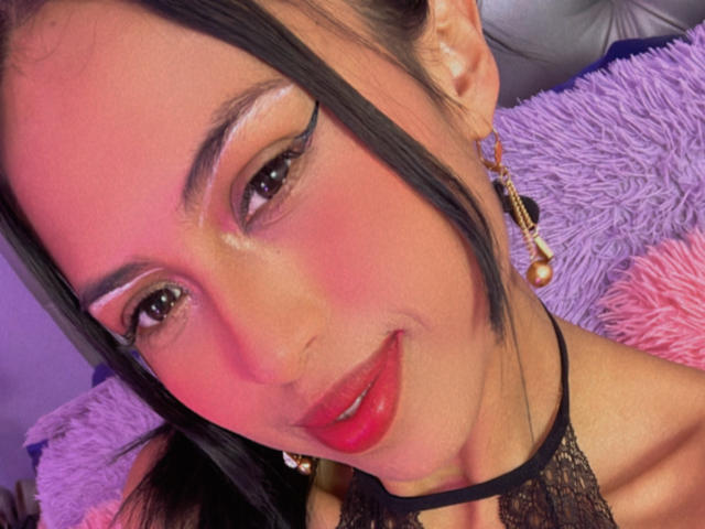 MyaaTyler's face, pink blushed cheeks, black eyeliner, red lipstick, smiling, black lace halter, long black hair, purple and pink fluffy pillows in background.