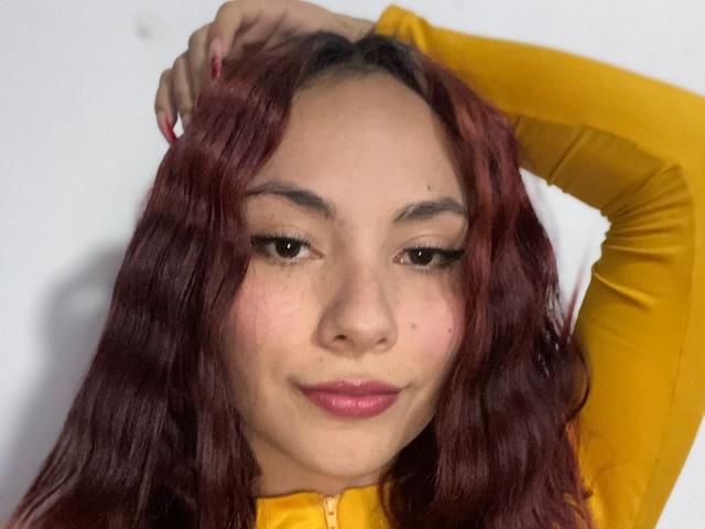 ValeryHarllow, con cabello rojo y ondulado, camisa amarilla de manga larga, una mano en el cabello, una sonrisa sutil, lápiz labial rosa y pestañas largas, mostrando su linda carita..