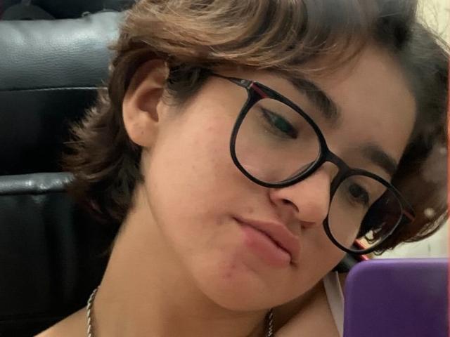BrianaSoica has short brown hair, black glasses, and is pouting with her tongue slightly out. She's wearing a necklace and is holding a purple phone.