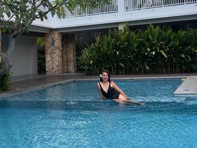 EmmaRose69, a woman with dark hair and a black one-piece swimsuit, sits in a blue pool, her tits and ass partially submerged, with green plants and a white building.