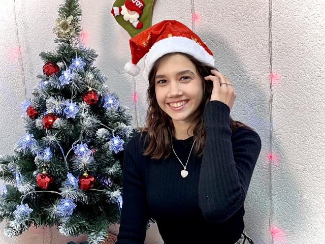 LettyCurly with brown hair, wearing a red Santa hat, black ribbed sweater, smiling, holding her hair, next to a decorated Christmas tree with red and blue ornaments.