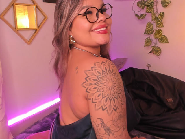 Juliana Sousa, con gli occhiali, sorridente, mostra un tatuaggio dettagliato di un mandala sulla spalla destra, indossa un top nero senza spalline, con luci rosa e viola, e c'è un cuscino nero sullo sfondo..