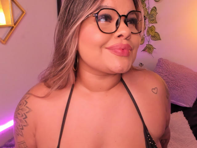 JulianaSoussa with glasses, pouty pink lips, blonde hair, black halter top, tattoos on shoulder and upper arm, heart tattoo, subtle makeup, pink lighting, slight smile.