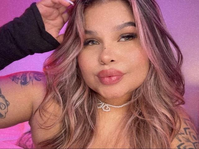 JulianaSoussa with pinkish-brown hair, full pink lips, a black sleeve, a diamond necklace, and tattoos on her arm, posing with a hand in her hair.