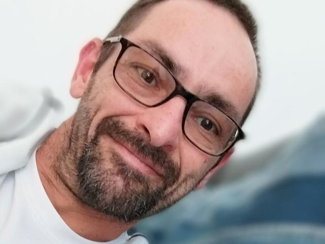 Ledjee has a balding head, dark glasses, and a graying beard, smiling slightly with a relaxed expression, wearing a white shirt. His nose is slightly crooked.