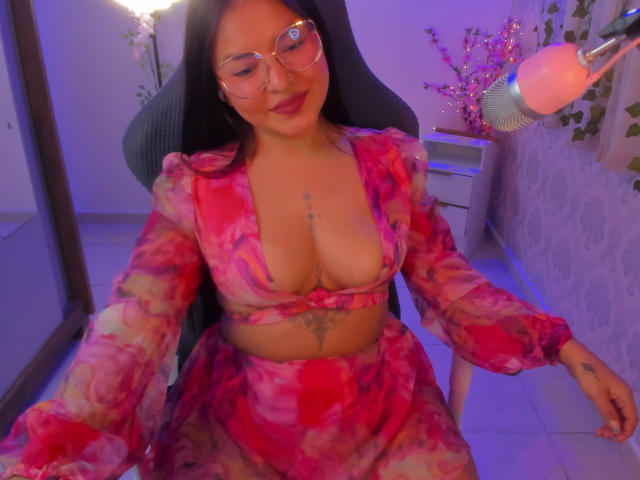 JazmineFerra with glasses, pink tie-dye top, and matching skirt, showing big tits and a tattoo below her tits, sitting in a chair with a pink ring light.