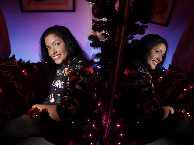 AnelisaReys with long black hair, smiling, wearing a festive sweater, black thigh-highs, and red ornament, reflected in a mirror, sitting in front of a Christmas tree.
