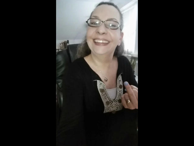 CashLadyVivian-hot indossa degli occhiali, ha i capelli castani raccolti in una coda di cavallo, sorride con un rossetto scuro, indossa una maglia nera con pizzo bianco, tiene una penna e sta seduta su una poltrona in pelle..