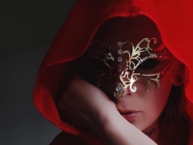 LizieLumiere with red hood, gold ornate mask, hand covering lower face, dark eyes, red light, and subtle smile. Her tits are partially visible under the hood.