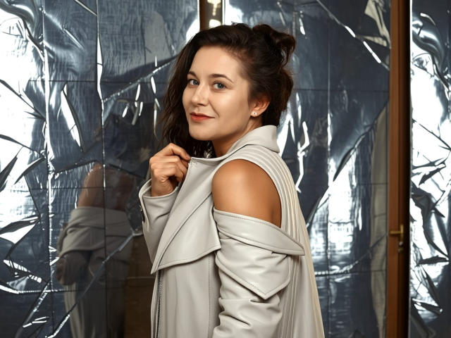 FlirtYolivia with brown hair, light skin, and red lipstick, wearing a grey, off-shoulder leather jacket, smiling slightly, holding jacket collar, reflected in shiny, silver backdrop.