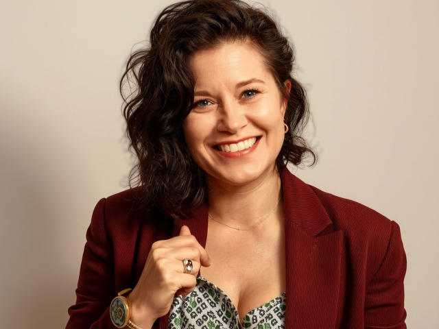 FlirtYolivia with wavy dark brown hair, smiling, wearing a red blazer, holding a patterned top, showing a ring, and a watch, with a cute, attractive face.