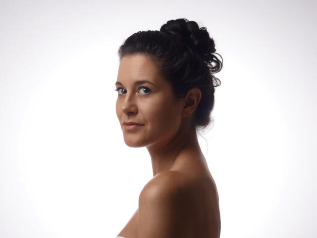 FlirtYolivia with dark hair in a messy bun, looking over her shoulder, smiling, topless, showing her bare shoulders and upper back, with a subtle, sexy, confident look.