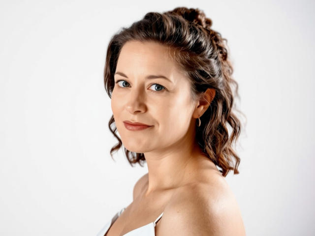 FlirtYolivia hat welliges, braunes Haar zu einem Pferdeschwanz gebunden, blaue Augen, helle Haut und ein leichtes Lächeln. Sie trägt ein weißes Top mit Trägern, das ihre nackten Schultern und ihre kleinen Brüste zeigt..
