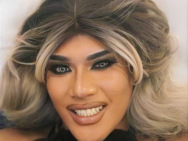 UrTsMidnightFantasy tem cabelo loiro ondulado com raízes escuras, sobrancelhas grossas, olhos verdes, delineador preto marcante e um sorriso que mostra dentes brancos. Os seios não estão visíveis..
