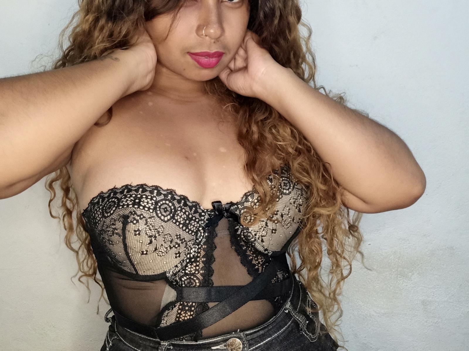 MiaAsly with curly brown hair, wearing a black lace strapless lingerie top, hands on her cheeks, pink lipstick, showing her big tits, against a plain white wall.