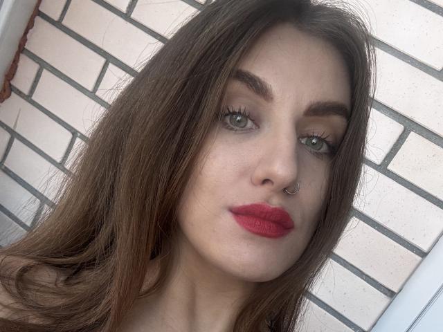 Kametia has long brown hair, green eyes, a nose ring, and red lipstick. Her tits are not visible. She's looking at the camera with a slight pout.