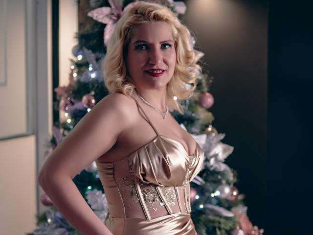 GiuliaAddams, com cabelos loiros e cacheados, vestindo um vestido dourado brilhante, exibindo seus seios grandes e sorrindo. Árvore de Natal com enfeites e luzes rosa ao fundo..