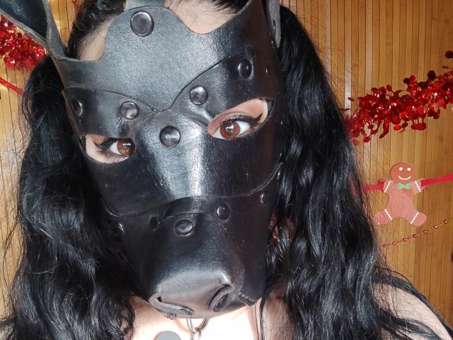 MiniBeast with black wavy hair wearing a black metal horse mask, showing brown eyes, with red tinsel and a gingerbread man in the background. Boobs barely visible.